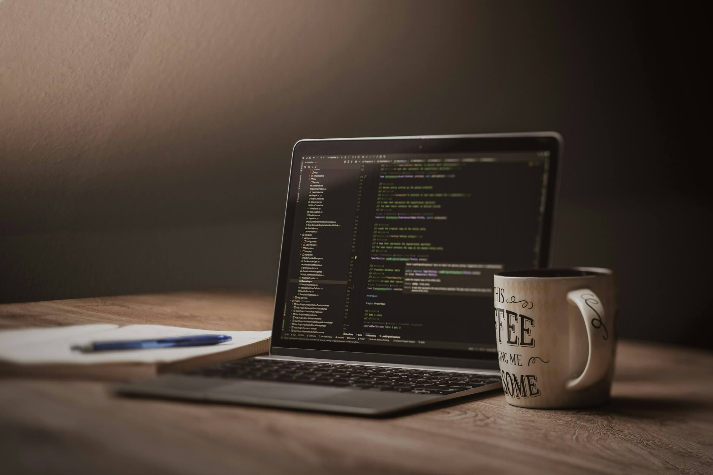 A laptop on a wooden desk with code on the screen, a notebook, and a coffee mug